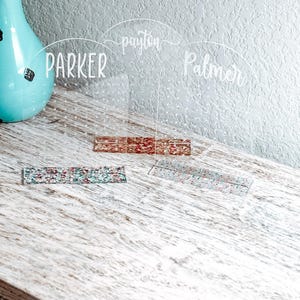 May include: Three clear acrylic table cards with names "Parker", "Payton", and "Palmer" written in white. The cards are on a light wood surface with glitter accents.