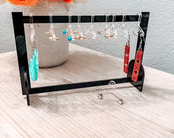 May include: A black acrylic earring stand with clear plastic hangers holding a variety of earrings. The earrings include turquoise and gold, red and gold, and clear crystal earrings.