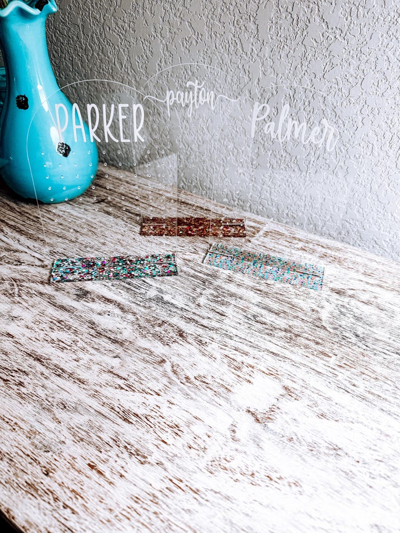 May include: Three clear acrylic earring holders with the names "Parker", "Payton", and "Palmer" written on them. Each holder has a row of small holes for earrings. The holders are sitting on a light wood surface with two colorful glitter strips in front of them.
