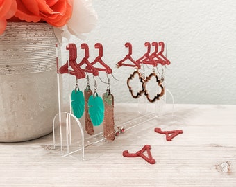 May include: A clear acrylic earring holder with red plastic hangers. The holder has multiple holes for earrings. There are several pairs of earrings on the hangers, including turquoise and gold leaf earrings.