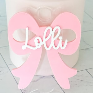 May include: A pink bow with a white cutout of the name "Lolli" in a cursive font.
