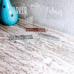 May include: Three clear acrylic earring holders with the names "Parker", "Payton", and "Palmer" written on them. Each holder has a row of small holes for earrings. The holders are sitting on a light wood surface with two colorful glitter strips in front of them.
