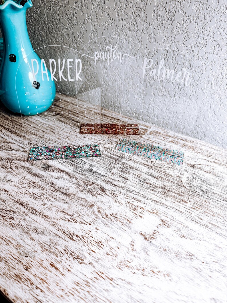 May include: Three clear acrylic earring holders with the names "Parker", "Payton", and "Palmer" written on them. Each holder has a row of small holes for earrings. The holders are sitting on a light wood surface with two colorful glitter strips in front of them.