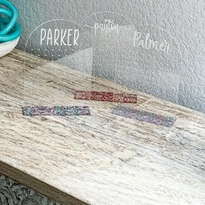May include: Two clear acrylic earring holders with the names "Parker", "Payton Palmer" written on them. Each holder has a row of holes for earrings and a decorative strip of fabric at the bottom.