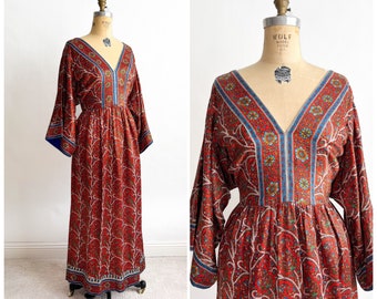 May include: A vintage red maxi dress with a V-neck and long, flared sleeves. The dress features a floral pattern with blue and white accents along the neckline, sleeves, and hem. The dress is displayed on a dress form.