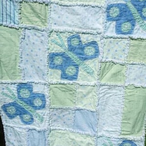 May include: A handmade patchwork quilt featuring a repeating butterfly design. The quilt is composed of various fabric squares in shades of blue, green, and white, with a frayed edge detail. The butterflies are appliqued in blue.