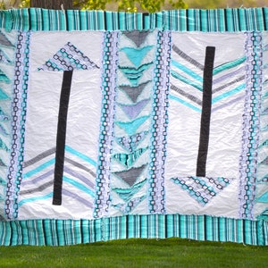 May include: A handmade quilt with a white background and turquoise, black, and gray arrow designs. The quilt features a striped border and decorative triangle accents. The quilt is displayed outdoors on a white fence.