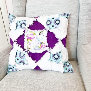 Op de afbeelding: Vierkant decoratief kussen met een patchwork ontwerp. Het kussen heeft witte, paarse en turquoise stoffen. Het middenpaneel heeft een bloemen- en vogelkooi print. Het kussen ligt op een grijze fauteuil.