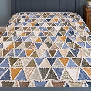 Puede incluir: Una colcha con un patrón repetitivo de triángulos en tonos azul, verde oliva, beige y amarillo mostaza. Las piezas de tela están unidas con costuras blancas deshilachadas y expuestas, creando una apariencia texturizada de rejilla.