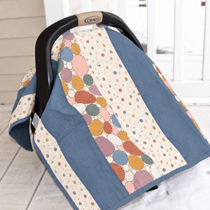 May include: A Graco infant car seat covered by a fabric canopy featuring vertical panels of solid dusty blue, a cream background with small multicolored dots, and a central section displaying a pattern of overlapping circles in muted tones.