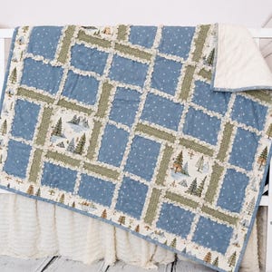 May include: A rag quilt featuring a patchwork design of blue fabric with white stars and cream fabric with a forest print of pine trees and mountains. The quilt has frayed seams and a solid cream-colored backing, draped over a white crib.