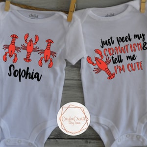 May include: Two white baby onesies with crawfish designs. One onesie has three red crawfish and the text "Sophia". The other onesie has one red crawfish and the text "just peel my crawfish & tell me I'm cute".