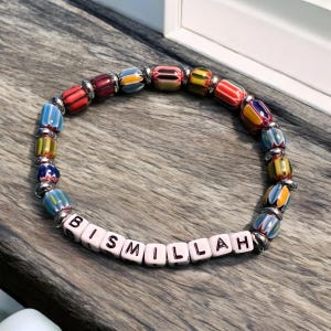 Pode incluir: Uma pulseira de contas com contas de vidro coloridas e detalhes prateados. A pulseira apresenta a palavra "BISMILLAH" escrita em contas de letras brancas. As contas são em tons de vermelho, azul, amarelo e laranja.