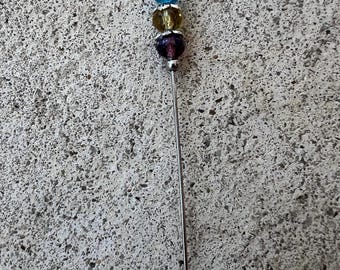May include: A silver pin with three glass beads. The beads are blue, yellow, and purple.