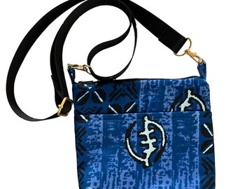 Puede incluir: Bolso bandolera azul con correa negra ajustable y herrajes dorados. El bolso presenta un estampado geométrico en blanco y negro sobre fondo azul. Tiene un bolsillo frontal y cierre de cremallera.