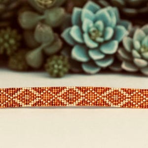 May include: A beaded bracelet with a geometric pattern in shades of orange, brown, and cream. The bracelet has a silver clasp and chain. The background features green and blue succulents.