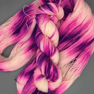 May include: A skein of hand-dyed yarn in shades of white, pink, and purple. The yarn is arranged in a loose coil with a few strands pulled out to show the color variations.