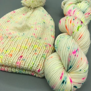 Puede incluir: Un gorro de punto blanco con un pomp&oacute;n en la parte superior y una madeja de hilo blanco con motas multicolores.