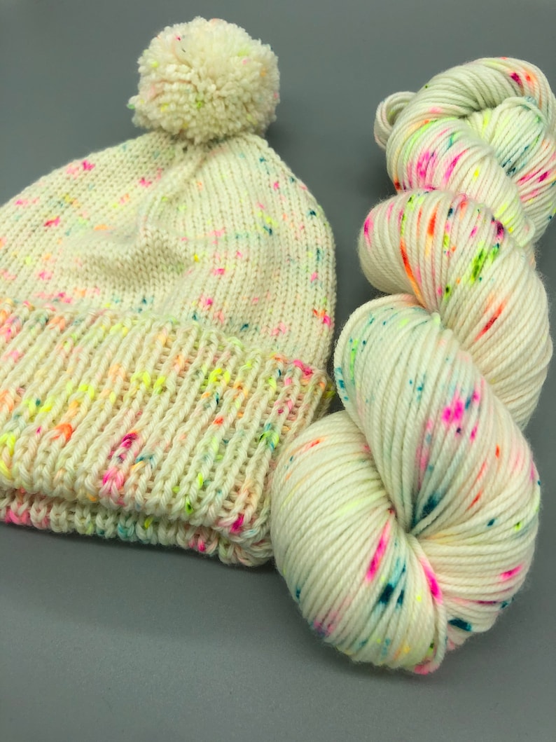 Puede incluir: Un gorro de punto blanco con un pomp&oacute;n en la parte superior y una madeja de hilo blanco con motas multicolores.