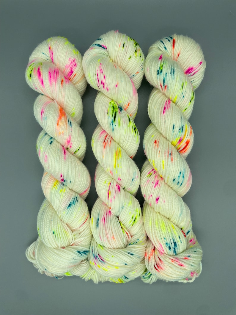 Puede incluir: Tres ovillos de hilo blanco con motas de colores. El hilo es una mezcla de blanco, rosa, naranja, amarillo, verde, azul y turquesa.