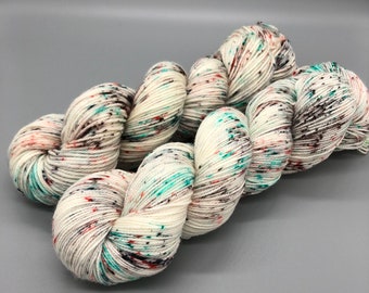 Puede incluir: Un ovillo de hilo blanco con salpicaduras de color turquesa, naranja y marrón. El hilo está dispuesto en una bobina suelta.