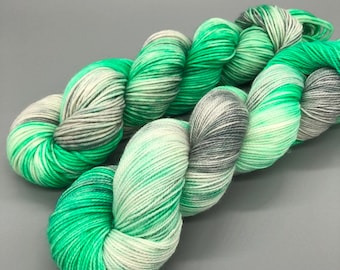 Puede incluir: Un ovillo de hilo en tonos de verde, blanco y gris. El hilo está teñido a mano y tiene un patrón de color variado.