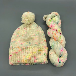 Puede incluir: Un gorro blanco de punto con un pomp&oacute;n y motas de colores, junto con un ovillo de lana blanca con motas de colores.