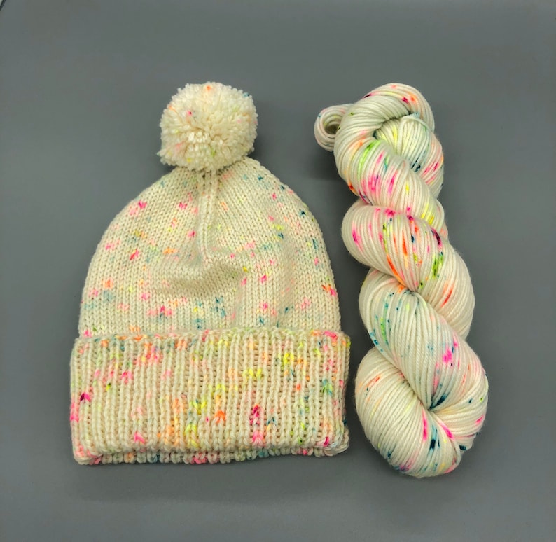Puede incluir: Un gorro blanco de punto con un pomp&oacute;n y motas de colores, junto con un ovillo de lana blanca con motas de colores.