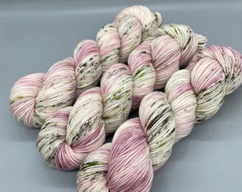 May include: Three skeins of hand-dyed yarn featuring a variegated color palette of soft pastel pink, creamy off-white, and ivory, accented with small, scattered speckles of dark brown and muted olive green throughout the twisted fiber strands.