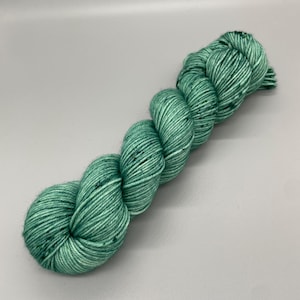 Könnte beinhalten: Ein Strang grüne Wolle mit weißen Flecken. Das Garn ist fest gewickelt und hat ein strukturiertes Aussehen. Die Farbe ist ein leuchtendes, helles Grün, geeignet für Strick- oder Häkelprojekte.