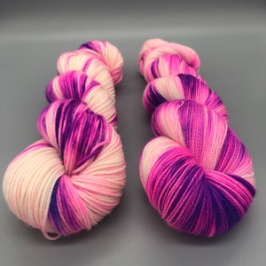 May include: Two skeins of yarn in a pink, white, and purple colorway. The yarn is a fingering weight and is perfect for knitting or crocheting.