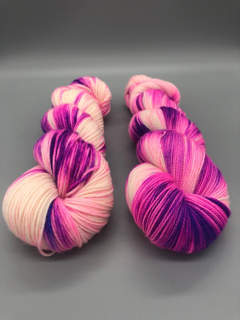 May include: Two skeins of yarn in a pink, white, and purple colorway. The yarn is a fingering weight and is perfect for knitting or crocheting.