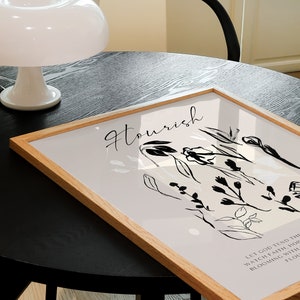May include: A framed print with a light grey background and black line drawings of flowers and leaves. The text "Flourish" is written in black cursive at the top of the print. The text "Let God tend the watch, faith, hope, blooming with a thousand flowers" is written in black at the bottom of the print.