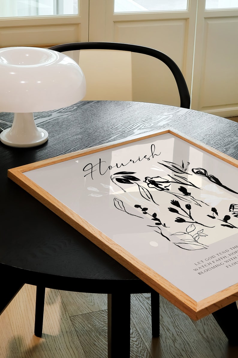 May include: A framed print with a light grey background and black line drawings of flowers and leaves. The text "Flourish" is written in black cursive at the top of the print. The text "Let God tend the watch, faith, hope, blooming with a thousand flowers" is written in black at the bottom of the print.