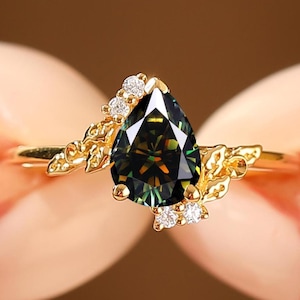Op de afbeelding: Een gouden ring met een peervormige, donkergroene edelsteen. De ring is versierd met kleine, ronde, heldere edelstenen en bladeraccenten. De ring wordt tussen twee vingers gehouden.