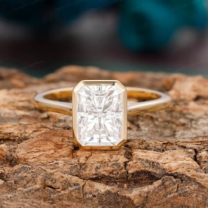 Pode incluir: Um anel de noivado de ouro com um grande diamante retangular transparente. O anel está colocado sobre uma superfície de madeira.