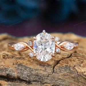 May include: Rose gold oval-cut diamond engagement ring with twisted band and marquise-shaped side stones. The center stone is a large, brilliant oval diamond, surrounded by smaller diamonds. The band is a delicate twisted design, with two marquise-shaped diamonds set on either side of the center stone. The ring is set on a rustic wooden surface.