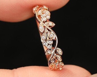 May include: Rose gold ring with a floral design. The ring features small, clear gemstones and a delicate leaf pattern. The ring is held between two fingers against a black background. The ring is a piece of jewelry.