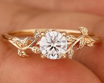 May include: A gold ring featuring a large, round, clear gemstone. The ring's band is adorned with delicate, leaf-like designs that extend around the central stone, creating a nature-inspired aesthetic. The ring is held up by a person's finger.