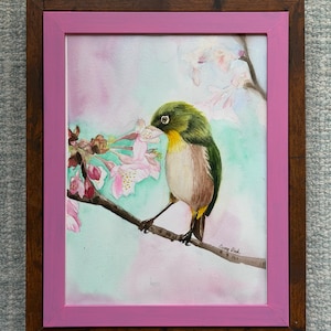 May include: Watercolor painting of a small bird perched on a branch with pink cherry blossoms. The bird has green, yellow, and brown feathers. The artwork is framed in a pink and brown wooden frame.
