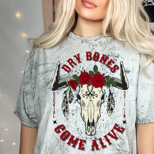 Könnte beinhalten: Weißes T-Shirt mit einem abgewetzten Look, das einen Schädel mit Hörnern und roten Rosen zeigt. Der Text "Dry Bones Come Alive" ist um den Schädel herum geschrieben, mit dem Text "Ezekial 37:1-10" unter dem Schädel.