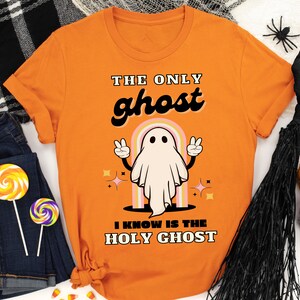 Puede incluir: Camiseta naranja con el texto "THE ONLY ghost I KNOW IS THE HOLY GHOST" y una ilustración de un fantasma de dibujos animados. El fantasma es blanco con brazos negros y hace signos de paz. La camiseta es parte de un tema de Halloween.