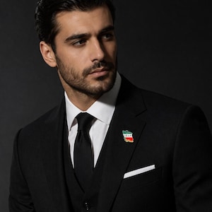 May include: A black suit with a white dress shirt and black tie. A small pin with the Iranian flag is on the lapel. The suit has a pocket square. The background is dark.