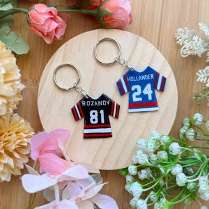 May include: Two jersey-shaped keychains. One is black with white and red stripes, the name "ROZANOV" and the number "81". The other is blue with white and red stripes, the name "HOLLANDER" and the number "24". Both have silver key rings.