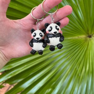 May include: Two panda bear keyrings. Each panda has a black and white body, black ears, and rosy cheeks. The keyrings are held by a gold-coloured ring. The background is a green palm leaf.