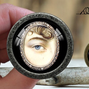 Peut inclure: Un bouton rond noir et argent avec un insert en verre représentant un portrait de l'œil d'une personne avec un iris bleu. L'insert en verre est entouré d'une bordure argentée décorative.