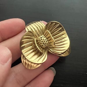 May include: A gold-colored flower-shaped brooch. The brooch has detailed petals and a textured center, resembling a dogwood flower. The brooch is made of metal and has a shiny finish.