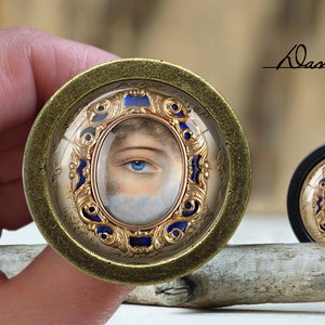 May include: A gold-framed, blue and white cameo with a single eye painted in the centre. The cameo is set in a round, brass-coloured frame.