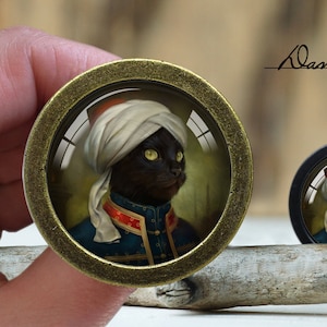 May include: A black cat wearing a white turban and a blue and red jacket. The cat is framed in a gold-coloured round pendant.