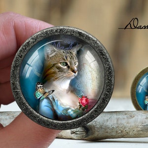 May include: A round, silver-toned, glass-covered, decorative knob with a picture of a cat wearing a blue dress and a butterfly on its shoulder. The cat has brown and white fur and green eyes. The background is blue and white with a floral pattern.